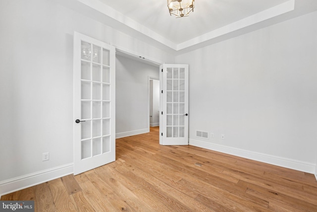 empty room with a chandelier, a raised ceiling, light wood-type flooring, and french doors