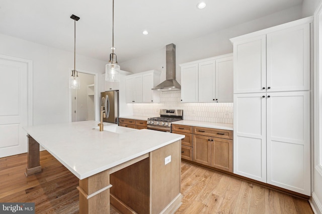 kitchen with wall chimney exhaust hood, hanging light fixtures, an island with sink, white cabinets, and appliances with stainless steel finishes