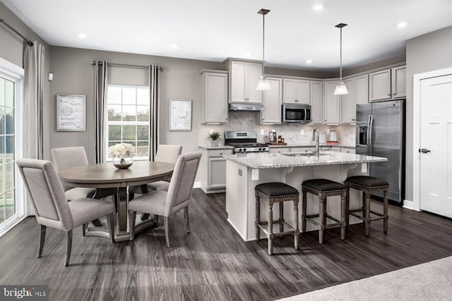 kitchen featuring a kitchen island with sink, hanging light fixtures, decorative backsplash, light stone countertops, and appliances with stainless steel finishes