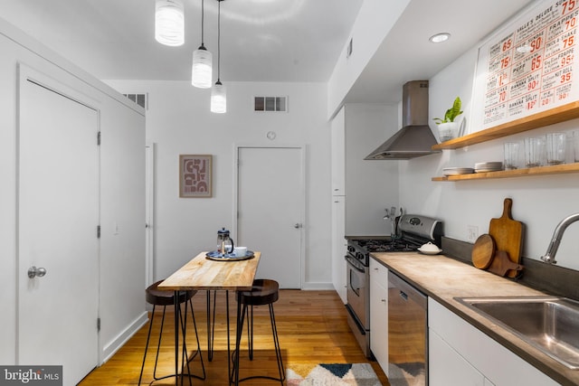 kitchen featuring wall chimney range hood, sink, light hardwood / wood-style flooring, decorative light fixtures, and stainless steel appliances