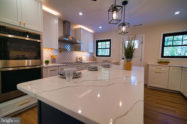 kitchen featuring light stone counters, wall chimney exhaust hood, hanging light fixtures, white cabinetry, and stainless steel double oven