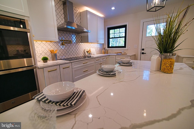 kitchen featuring black electric cooktop, stainless steel double oven, light stone counters, wall chimney exhaust hood, and white cabinets