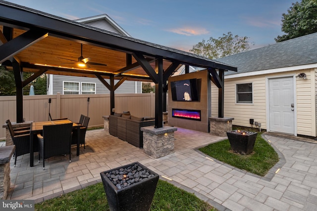 patio terrace at dusk with a gazebo, ceiling fan, and an outdoor living space with a fireplace