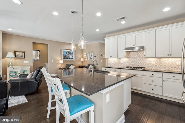 kitchen with sink, hanging light fixtures, dark stone countertops, stainless steel gas stovetop, and a center island with sink