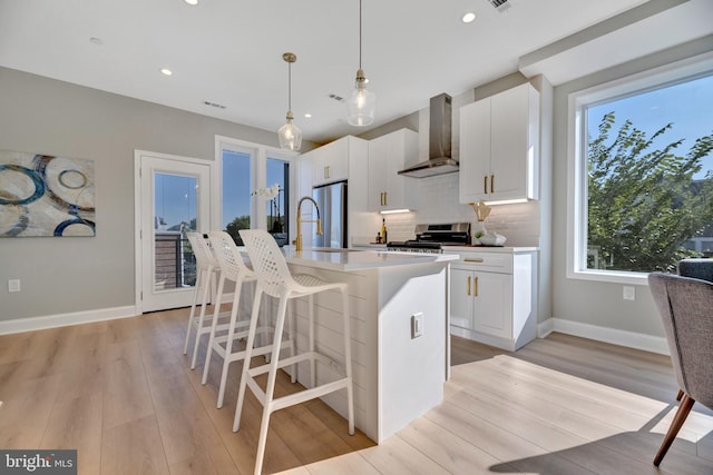 kitchen featuring wall chimney range hood, decorative light fixtures, a breakfast bar area, a kitchen island with sink, and white cabinets