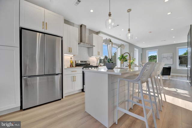 kitchen with appliances with stainless steel finishes, a kitchen island with sink, wall chimney range hood, white cabinetry, and hanging light fixtures