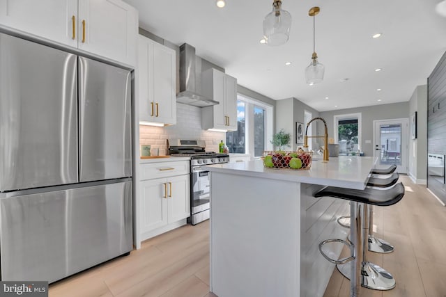 kitchen with appliances with stainless steel finishes, wall chimney range hood, decorative light fixtures, a center island with sink, and white cabinetry