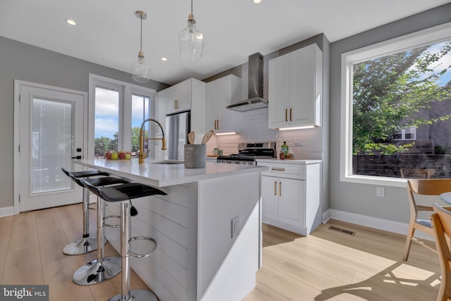 kitchen featuring appliances with stainless steel finishes, wall chimney range hood, pendant lighting, a center island with sink, and white cabinets