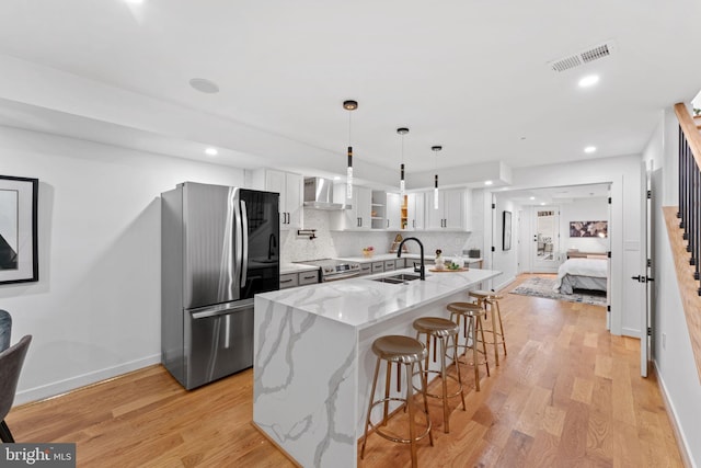 kitchen featuring appliances with stainless steel finishes, a kitchen island with sink, light stone countertops, white cabinets, and wall chimney exhaust hood