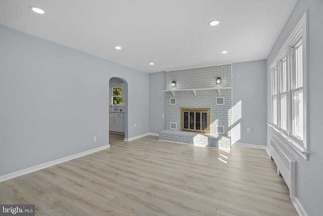 unfurnished living room with radiator heating unit, light wood-type flooring, a brick fireplace, and plenty of natural light