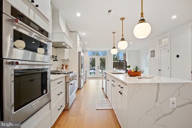 kitchen featuring custom range hood, stainless steel appliances, a center island with sink, white cabinets, and hanging light fixtures