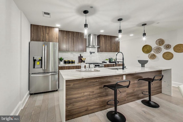 kitchen featuring sink, wall chimney exhaust hood, stainless steel appliances, light stone counters, and pendant lighting