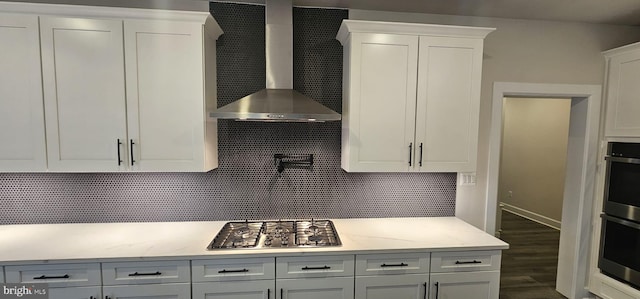 kitchen featuring wall chimney range hood, appliances with stainless steel finishes, white cabinetry, light stone counters, and decorative backsplash