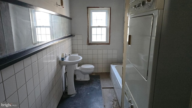 bathroom featuring a bathing tub, toilet, tile walls, and washer / dryer