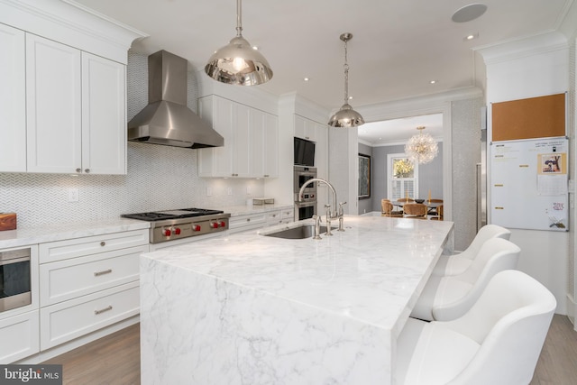 kitchen featuring wall chimney range hood, a center island with sink, ornamental molding, wood finished floors, and stainless steel appliances