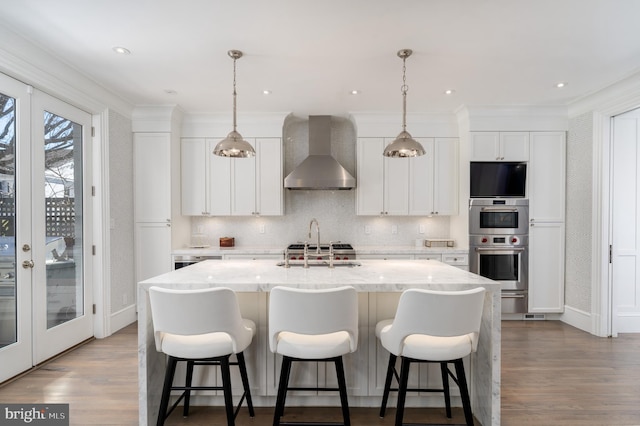 kitchen with stainless steel double oven, a center island with sink, wall chimney exhaust hood, and wood finished floors