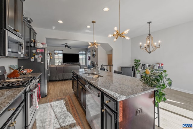 kitchen with light stone counters, ceiling fan with notable chandelier, stainless steel appliances, sink, and a center island with sink