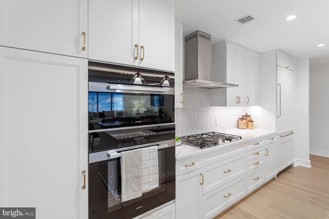 kitchen with stainless steel gas stovetop, wall chimney range hood, double oven, light stone counters, and white cabinetry