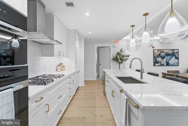 kitchen featuring a kitchen island with sink, wall chimney range hood, sink, hanging light fixtures, and white cabinetry