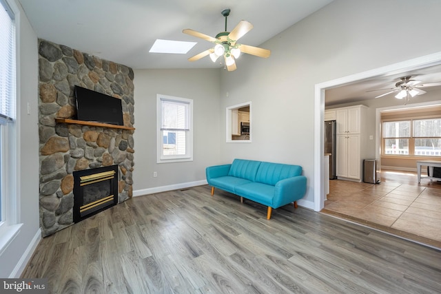 sitting room with ceiling fan, a stone fireplace, vaulted ceiling with skylight, and light wood-type flooring