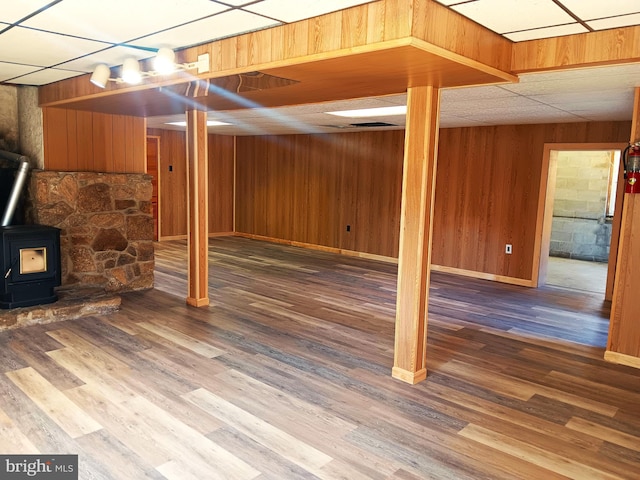 basement featuring a wood stove, wooden walls, a drop ceiling, and wood-type flooring