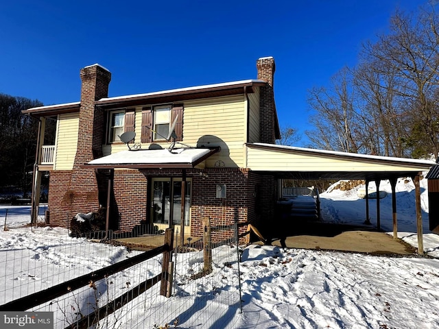 snow covered rear of property featuring a carport, brick siding, fence, and a chimney