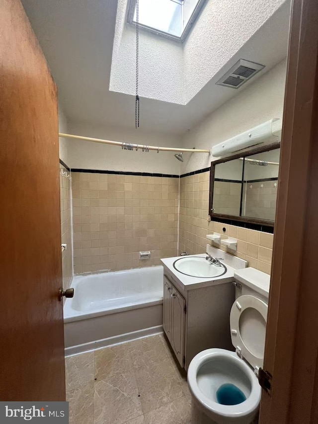 full bathroom featuring a skylight, tiled shower / bath combo, toilet, vanity, and tile walls