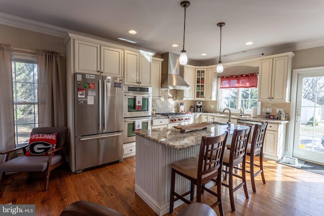 kitchen featuring light stone counters, hanging light fixtures, a kitchen island, wall chimney range hood, and appliances with stainless steel finishes