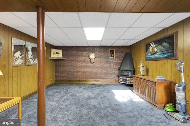 finished basement with wooden walls, brick wall, a drop ceiling, carpet floors, and a wood stove