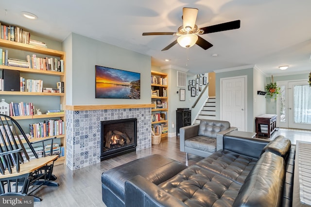 living room featuring a tile fireplace, built in features, wood-type flooring, ornamental molding, and ceiling fan