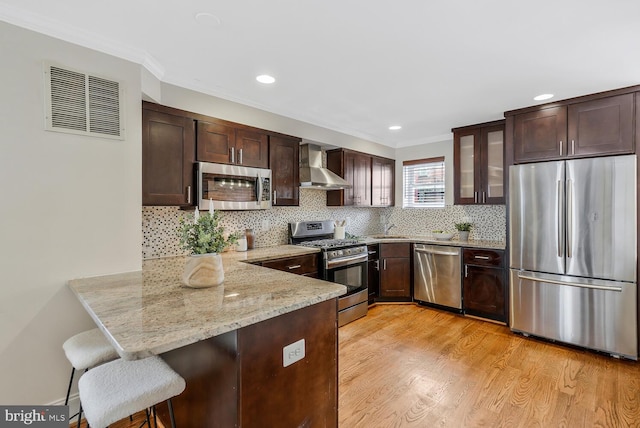 kitchen featuring dark brown cabinetry, wall chimney exhaust hood, stainless steel appliances, kitchen peninsula, and a breakfast bar area