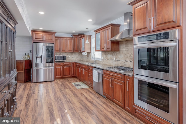 kitchen with wall chimney range hood, sink, crown molding, light stone countertops, and stainless steel appliances