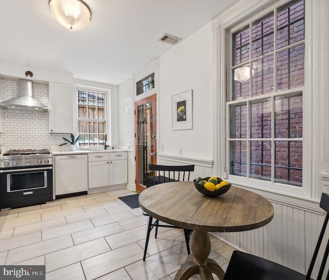 kitchen featuring gas range, white dishwasher, white cabinets, tasteful backsplash, and wall chimney range hood