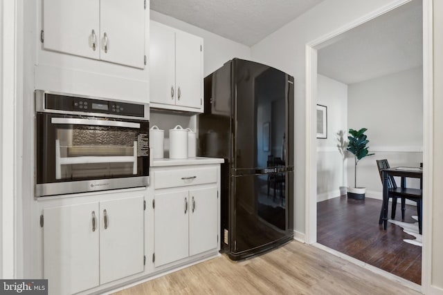 kitchen with white cabinets, stainless steel oven, a textured ceiling, black fridge, and light wood-type flooring