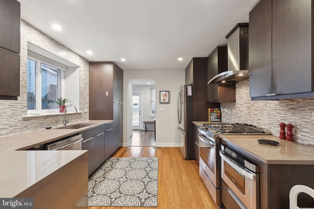 kitchen with sink, wall chimney range hood, light hardwood / wood-style flooring, decorative backsplash, and appliances with stainless steel finishes