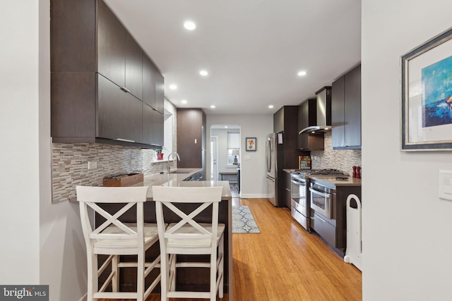 kitchen with wall chimney range hood, backsplash, kitchen peninsula, a breakfast bar, and appliances with stainless steel finishes