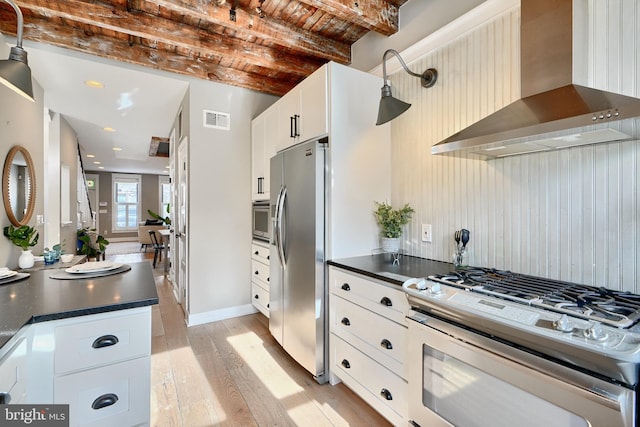 kitchen featuring stainless steel appliances, wall chimney range hood, light hardwood / wood-style flooring, beamed ceiling, and white cabinetry