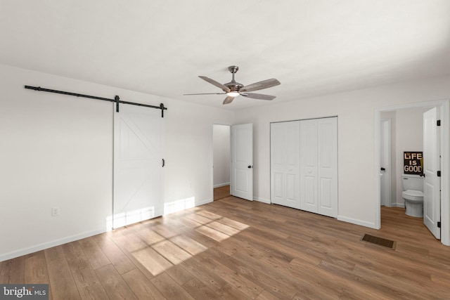 unfurnished bedroom with ensuite bathroom, ceiling fan, hardwood / wood-style flooring, and a barn door