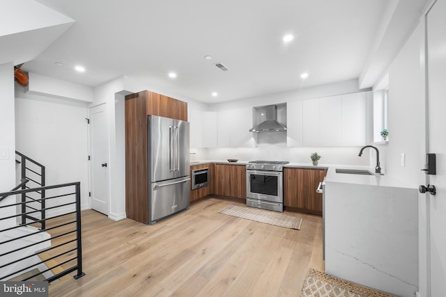 kitchen with white cabinetry, sink, wall chimney range hood, light hardwood / wood-style flooring, and appliances with stainless steel finishes