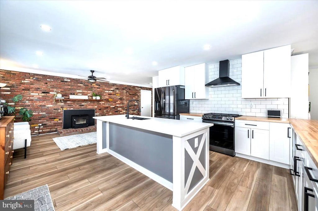 kitchen with black fridge with ice dispenser, wall chimney range hood, gas stove, white cabinets, and sink