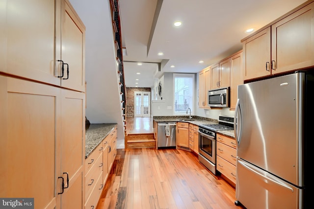 kitchen with light wood-style flooring, appliances with stainless steel finishes, light brown cabinets, a sink, and dark stone counters