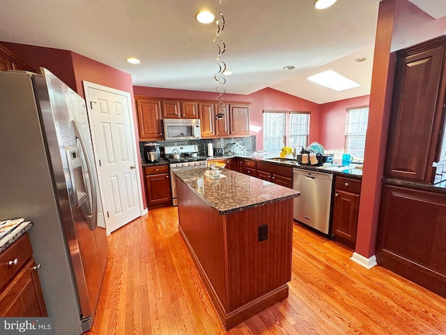 kitchen with stainless steel appliances, a kitchen island, light wood finished floors, lofted ceiling with skylight, and dark stone countertops