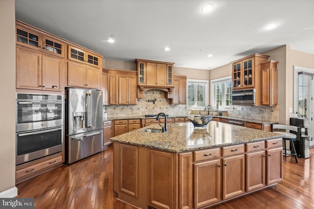 kitchen featuring dark wood-style floors, a center island with sink, a sink, appliances with stainless steel finishes, and tasteful backsplash