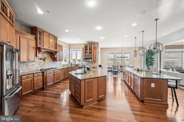 kitchen with brown cabinets, a center island with sink, a sink, stainless steel appliances, and an inviting chandelier