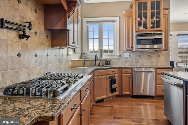 kitchen featuring brown cabinetry, wood finished floors, glass insert cabinets, appliances with stainless steel finishes, and backsplash