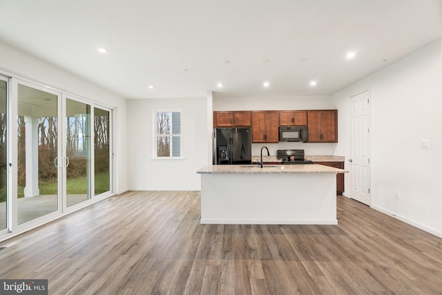 kitchen with black appliances, a center island with sink, sink, light stone counters, and wood-type flooring