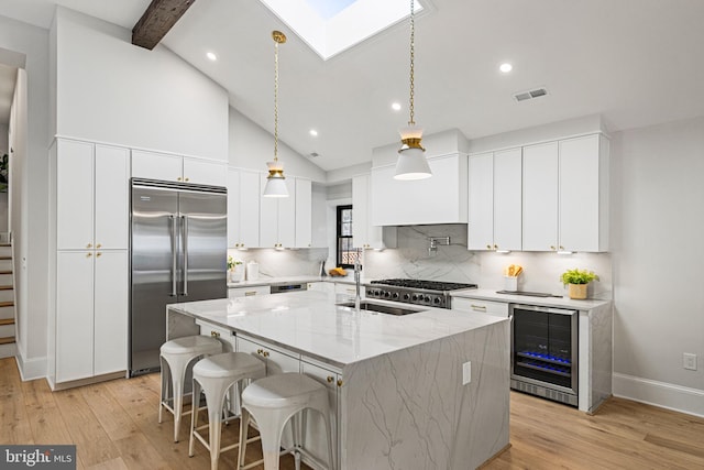 kitchen with wine cooler, appliances with stainless steel finishes, an island with sink, light stone counters, and white cabinets