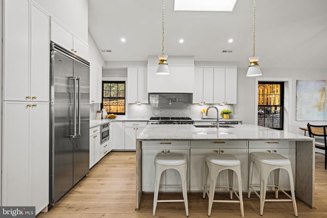 kitchen featuring an island with sink, built in appliances, sink, white cabinetry, and decorative light fixtures