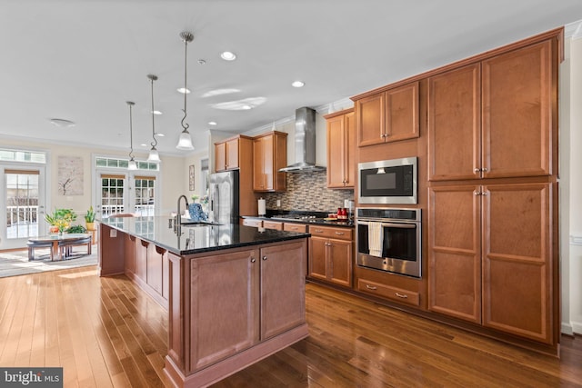 kitchen with pendant lighting, wall chimney range hood, stainless steel appliances, sink, and a center island with sink