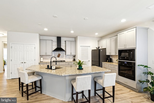 kitchen with sink, a breakfast bar area, black appliances, an island with sink, and wall chimney exhaust hood